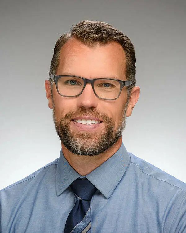 David Machiela is facing the camera smiling, in a professional pose. He wears glasses and is dressed smartly in a shirt and tie.