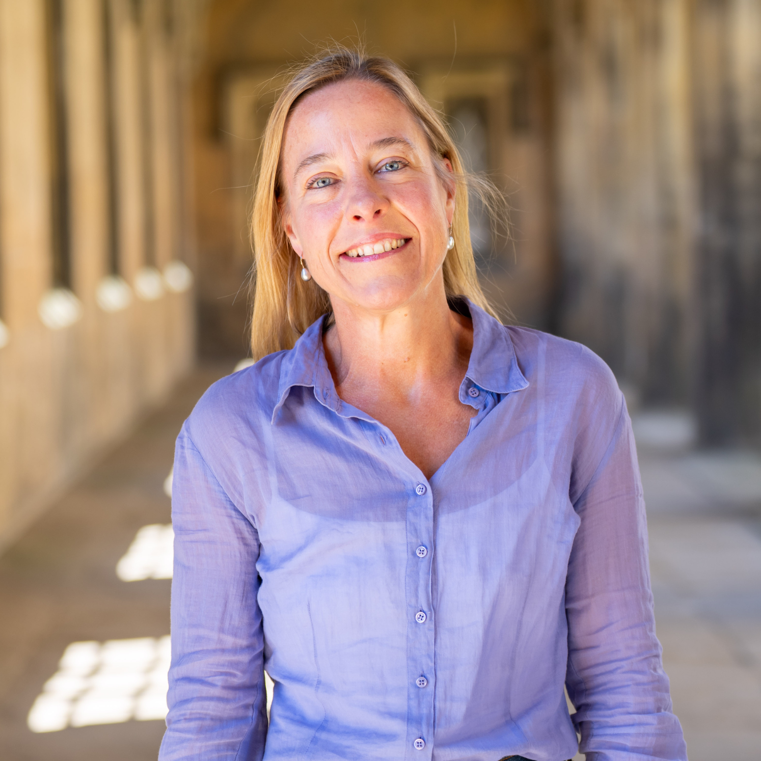 Dr Theresa Biberauer stands in New Court cloisters wearing a purple shirt, smiling at the camera