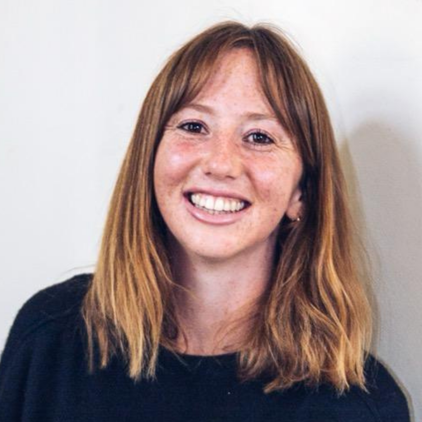 Dr Gillis is wearing a black jumper standing against a white wall, smiling at the camera