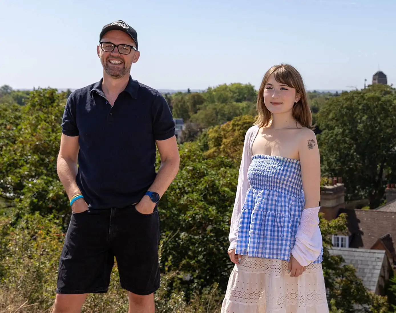 A man in a baseball cap and a young woman pose against a backdrop of the city of Cambridge