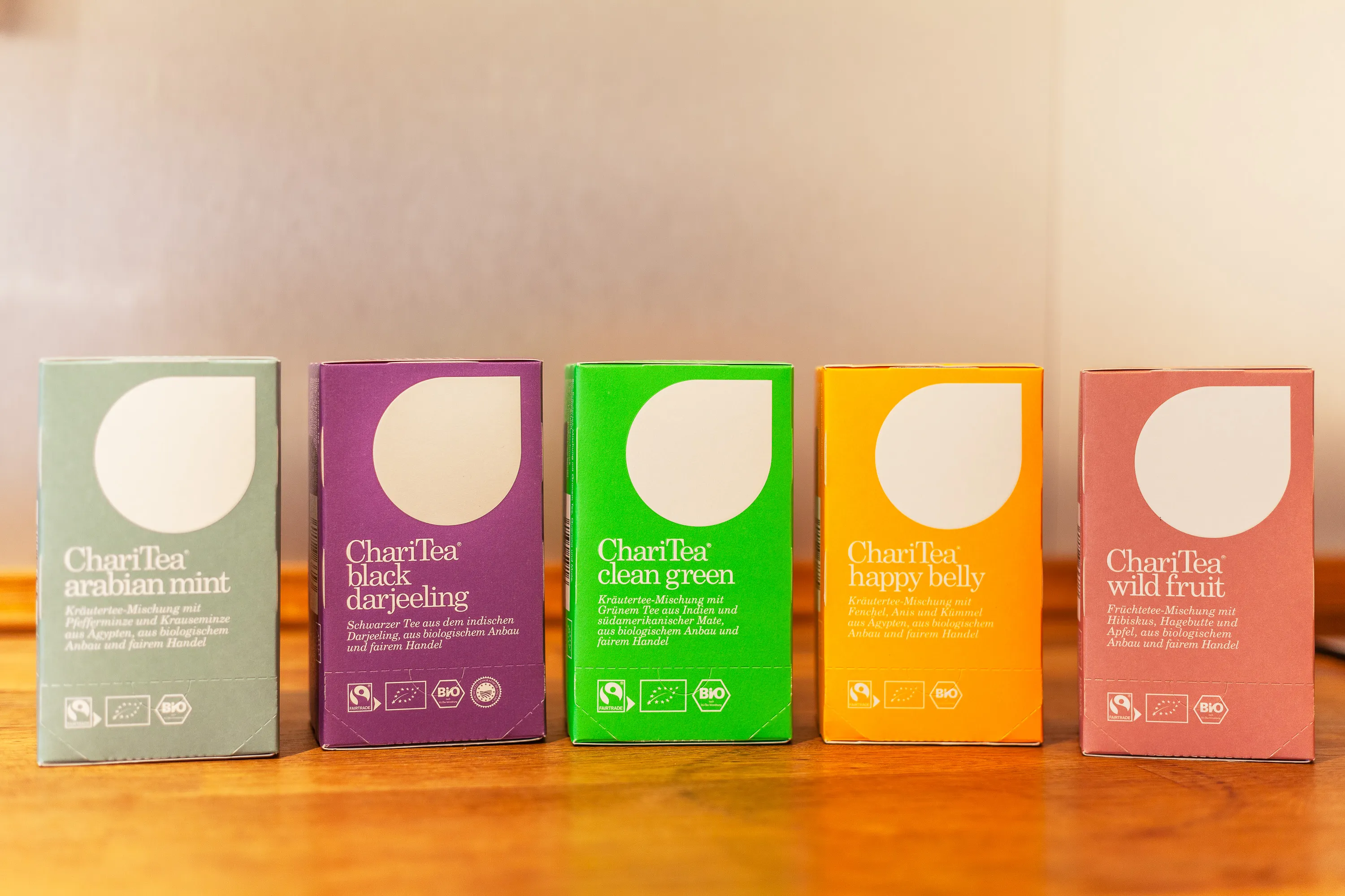 Fünf Tee-Packungen der Marke ChariTea steht auf der hölzernen Arbeitsplatte. Von links nach rechts zu sehen sind: Minze in mint, Darjeeling in lila, Grüntee in grün, Magen- und Darmtee in gelb und Fürchtetee in rot. Jeder dieser Tees ist bio und Fairtrade.