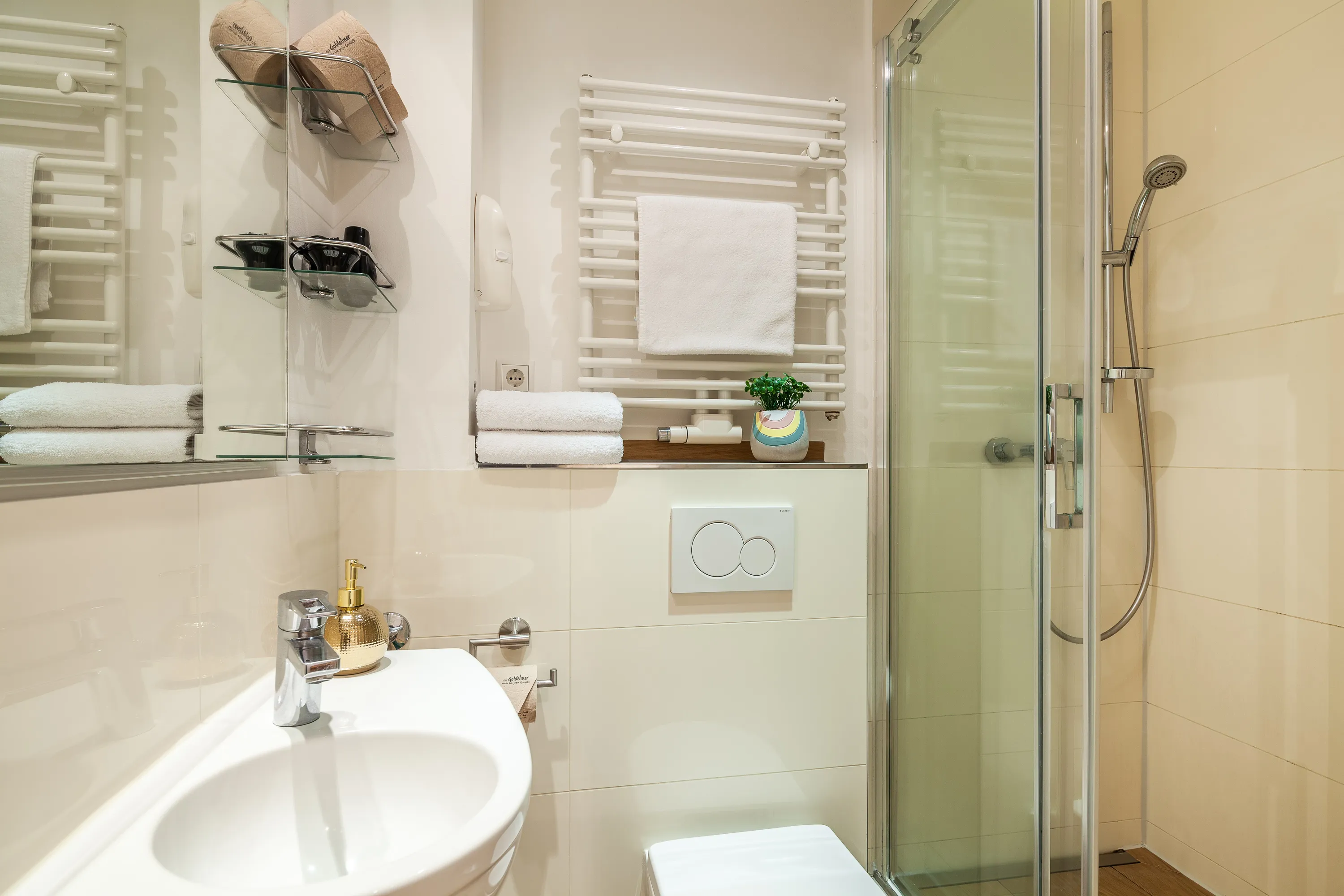 Das sanierte Badezimmer in Hamburg-Winterhude ist hell und in weiß und silber gehalten. Der Boden hat hellbraune Holzoptik. Links ist das kleine Waschbecken zu sehen, darüber ein großer Spiegel. Rechts daneben ist die Toilette und ein Handtuchhalter. Rechts ist die große Echtglasdusche, die durch eine Erhöhung begehbar ist. 
