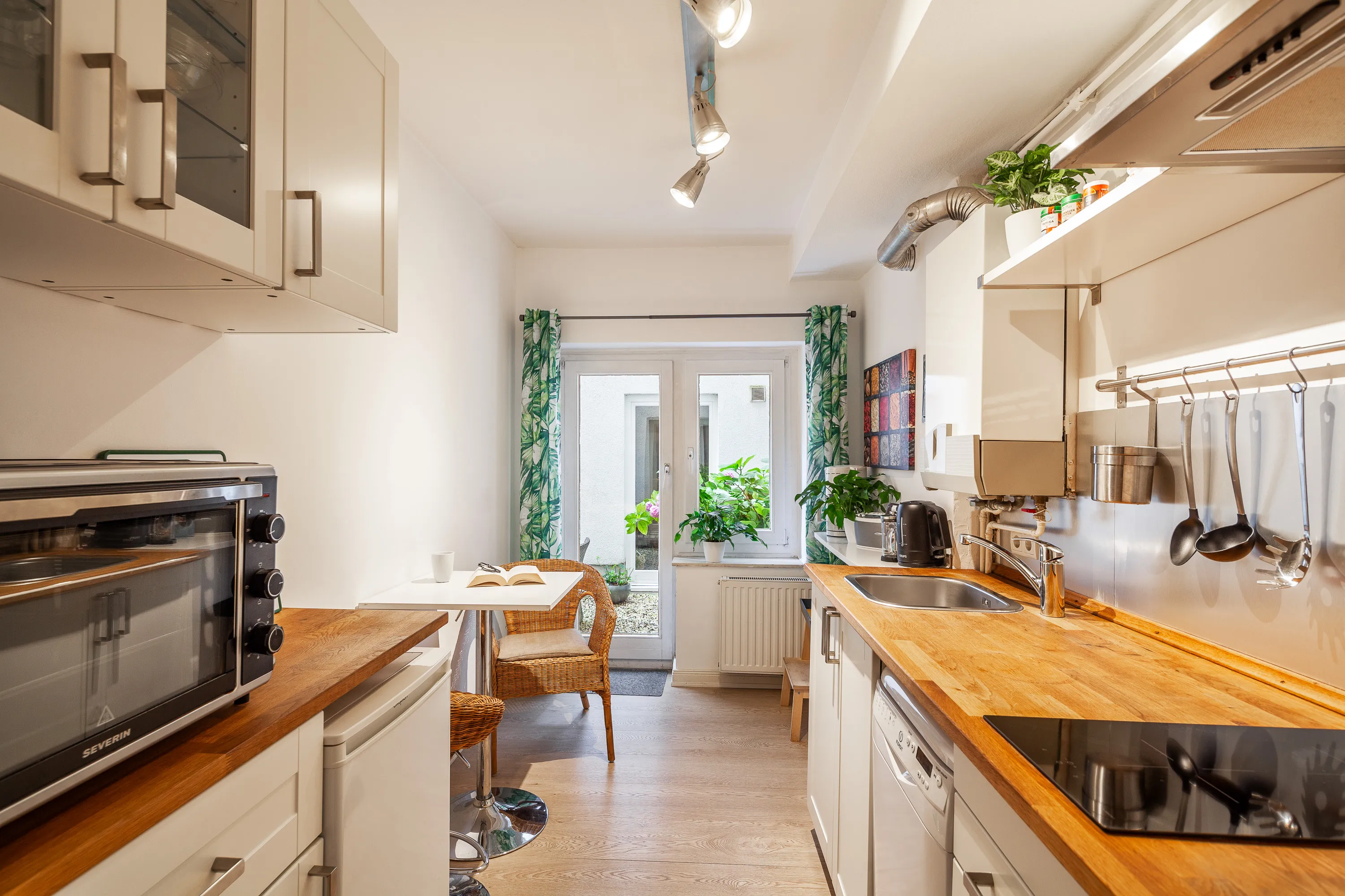 Die kleine Küche in der Ferienwohnung in Hamburg-Winterhude ist schmal und lang und hell. Am Eingang sind links und rechts Einbauschränke in weiß mit einer Holzarbeitsplatte. Links steht ein kleiner Backofen. Rechts ist das Kochfeld, hinter dem an der Wand Suppenkellen und Pfannenwender hängen. Im Hintergrund ist ein kleiner Esstisch zu sehen. Am Ende der Küche ist die große Fensterfront und die Tür, durch die es zur Terrasse geht. 