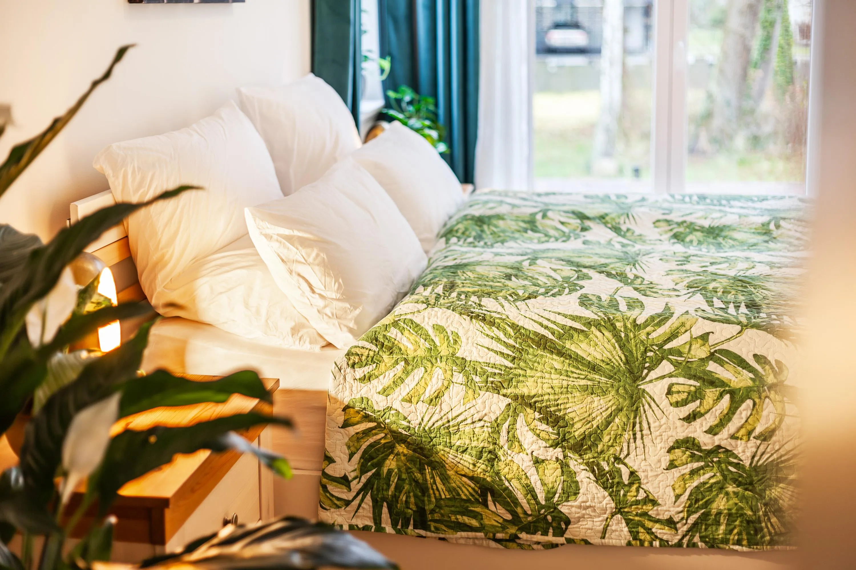 Gemütliches Bett mit weißen Kissen und Bettdecke mit grünem Blattmuster, umgeben von Zimmerpflanzen und natürlichem Licht
