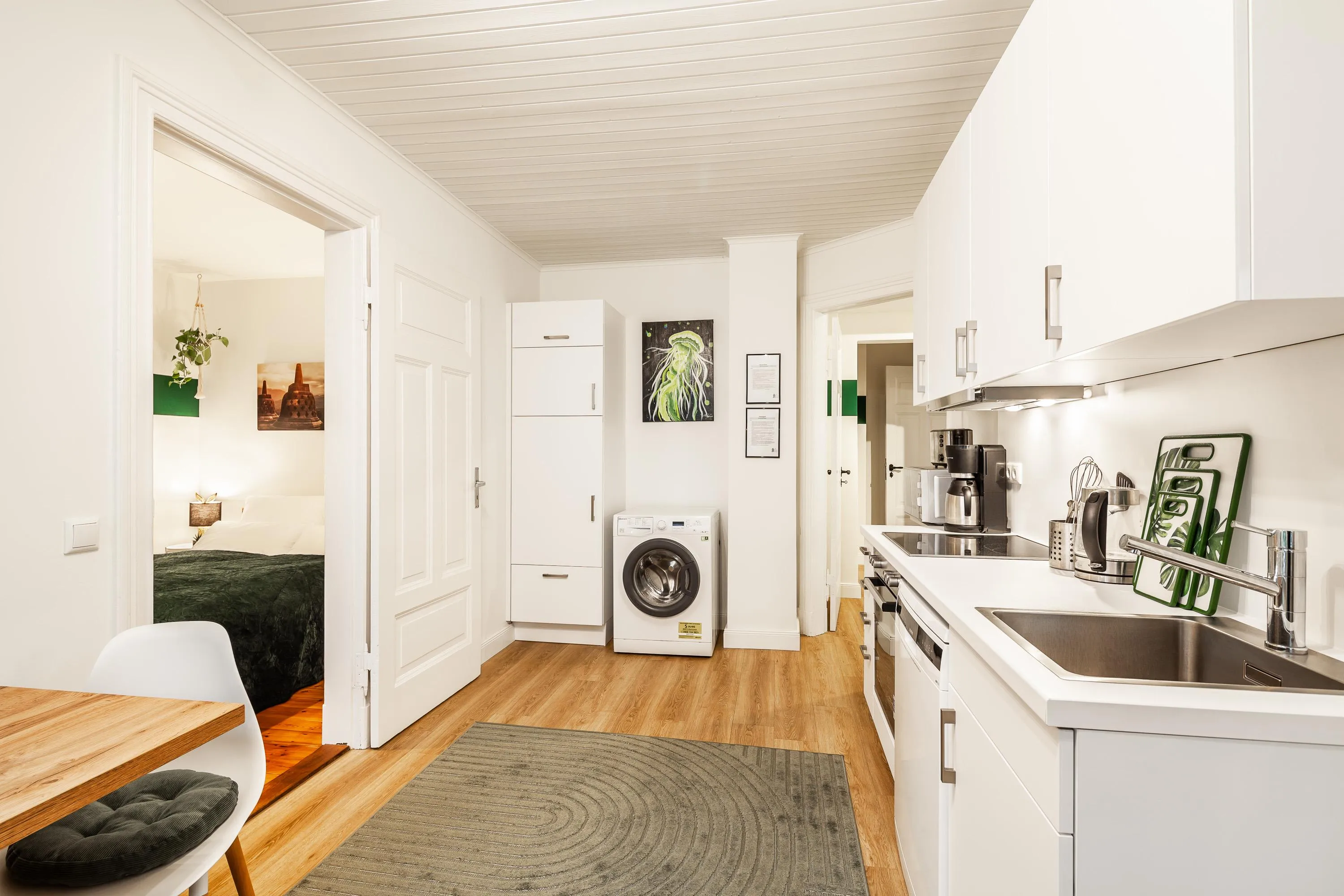 Helle Küche mit weißen Schränken, Waschmaschine, Spüle, Kaffeemaschine und Blick in angrenzendes Schlafzimmer mit grünem Bett