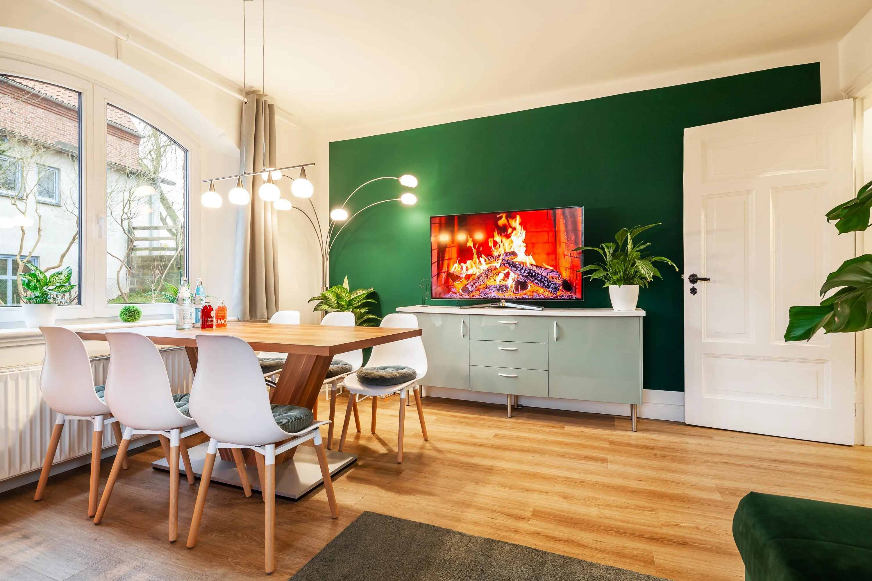 Esszimmer mit großem Holztisch, sechs weißen Stühlen, grün gestrichener Wand, Sideboard mit brennendem Kamin auf dem Fernseher und Pflanzen