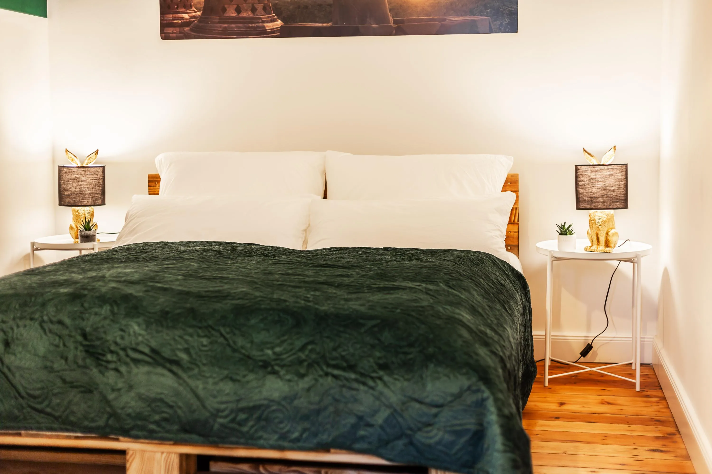 Doppelbett mit weißer Bettwäsche und dunkelgrünem Überwurf, zwei Nachttische mit Lampen und Dekoration in einem hellen Schlafzimmer