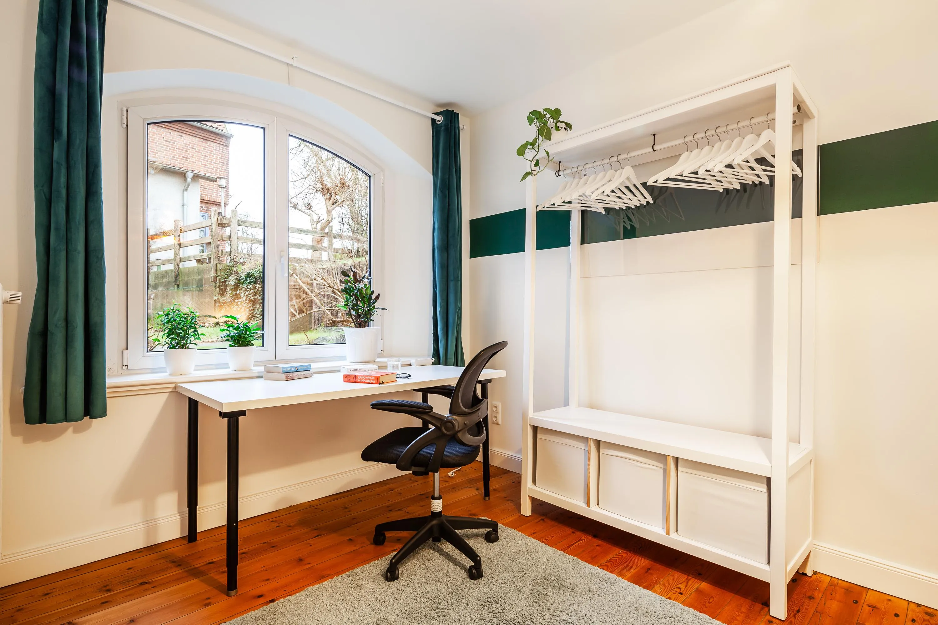 Helles Arbeitszimmer mit großem Fenster, Schreibtisch, schwarzem Bürostuhl und offenem weißem Kleiderregal mit Kleiderbügeln