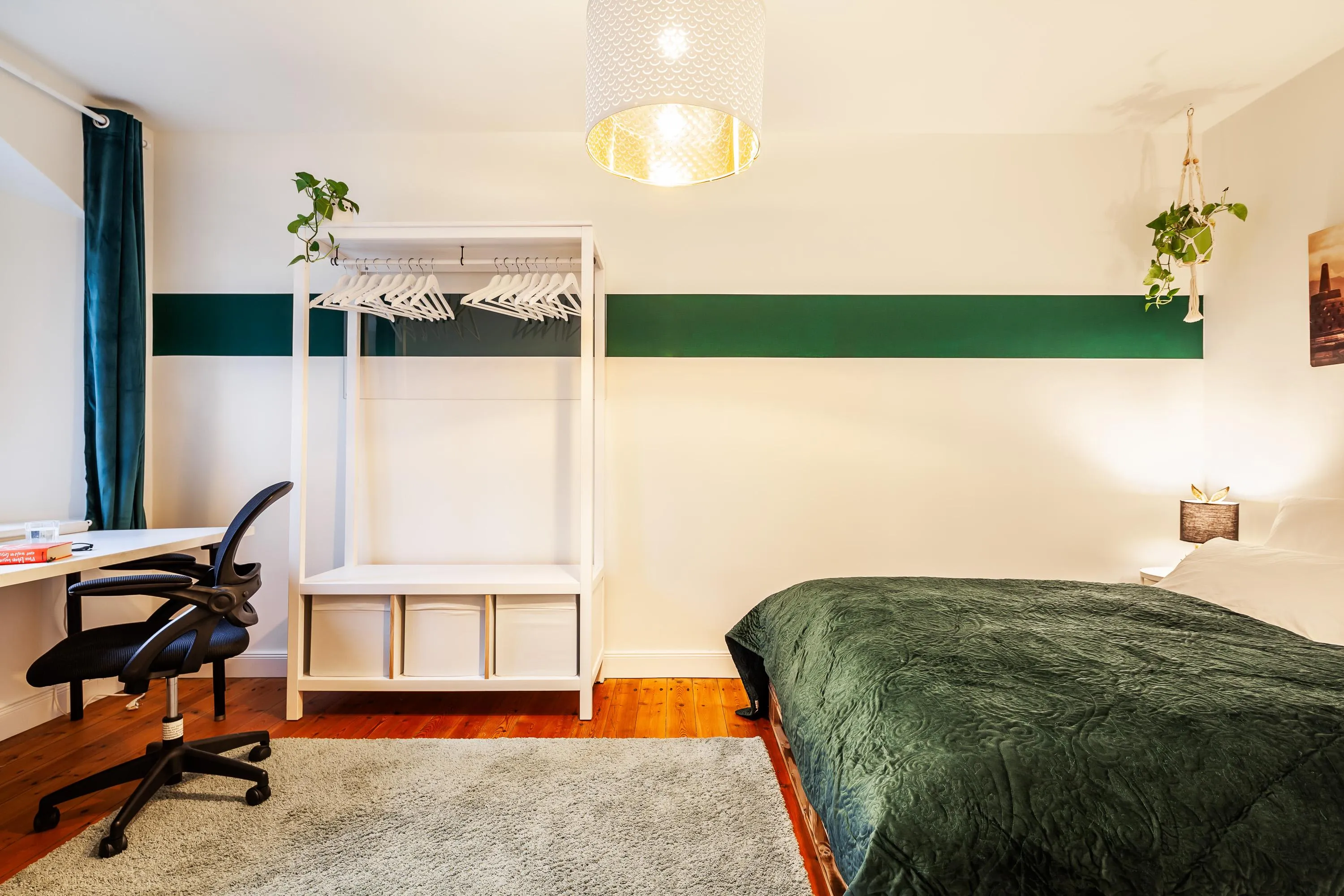 Schlafzimmer mit grünem Bettbezug, weißem Kleiderständer, Schreibtisch mit schwarzem Bürostuhl und grüner Wandstreifen