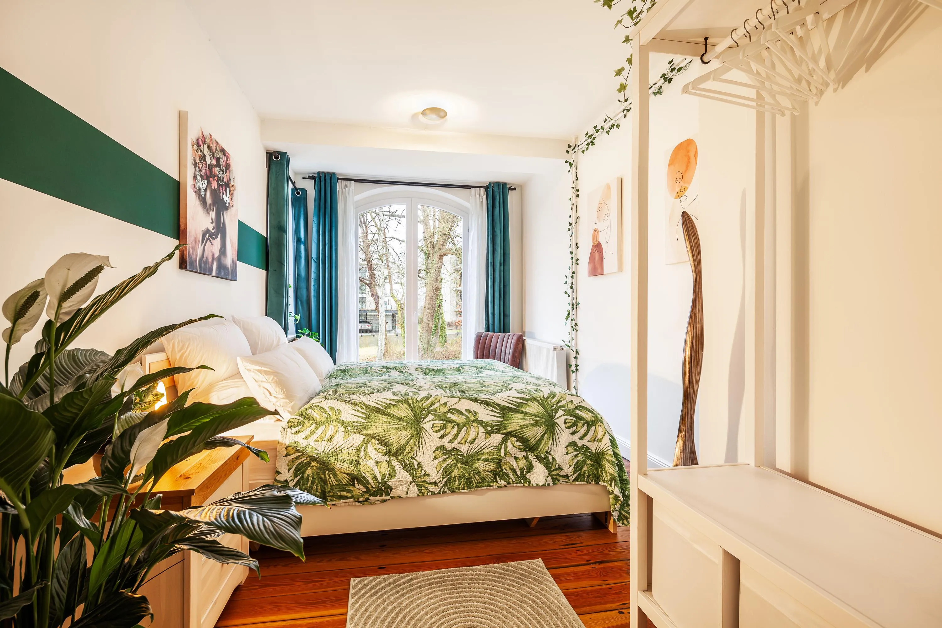 Helles Schlafzimmer mit großem Fenster, Bett mit grünem Blattmuster-Bettwäsche, Pflanzen und weißen Wänden mit Dekoration