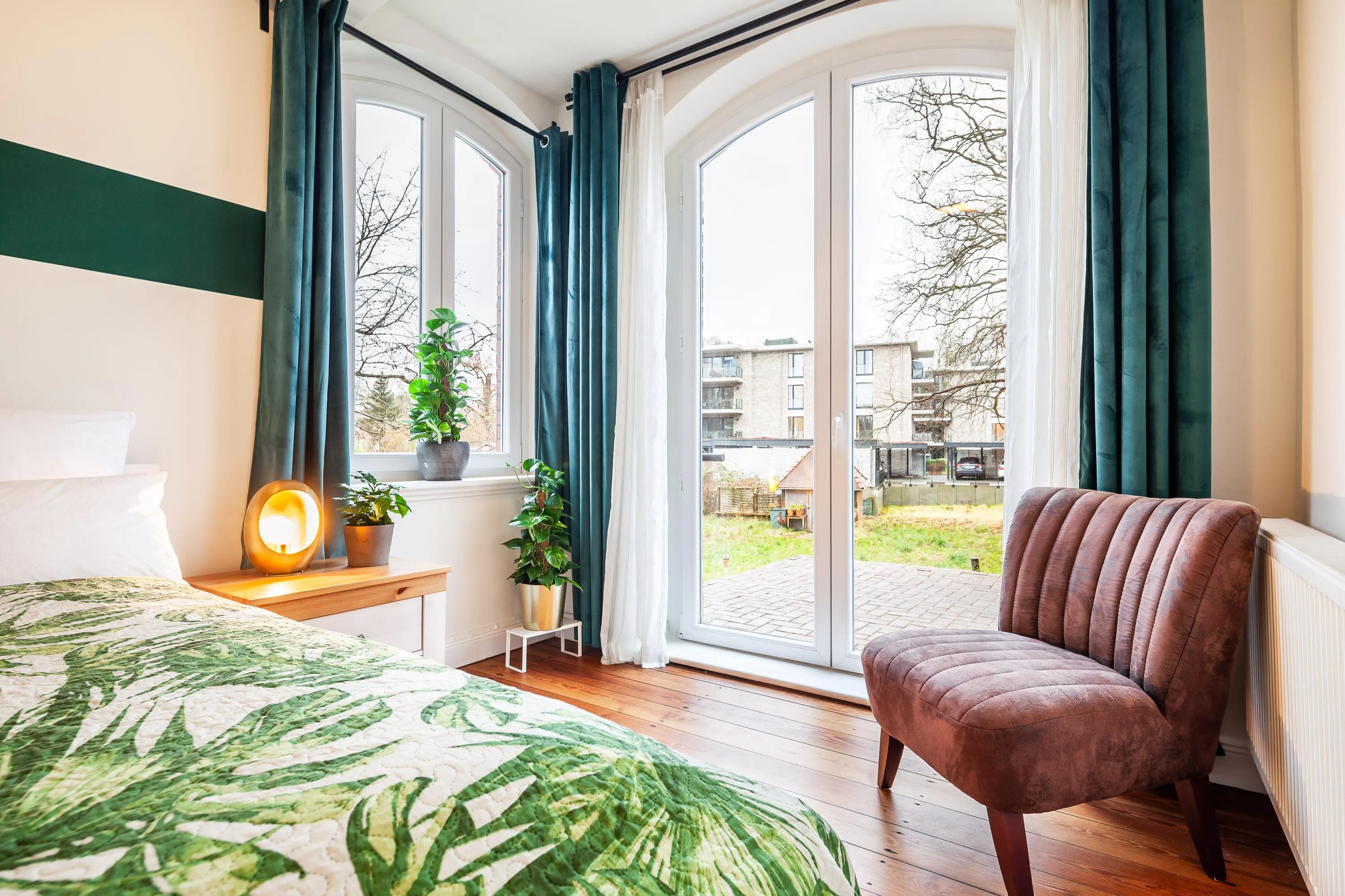 Helles Schlafzimmer mit großem Bogenfenster, grünen Vorhängen, Pflanzen auf Fensterbank, Bett mit grünem Blattmuster und braunem Sessel