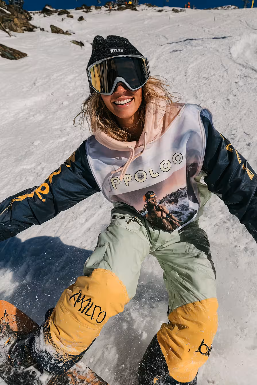 Smiling snowboarder wearing reflective goggles and colorful winter gear riding on snow-covered slope under clear blue sky.