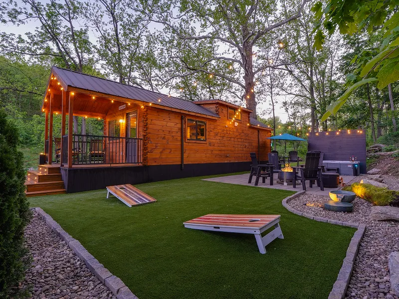 A wooden tiny house with string lights, a covered porch, artificial turf lawn with cornhole boards, outdoor seating area, and fire pit, surrounded by trees.