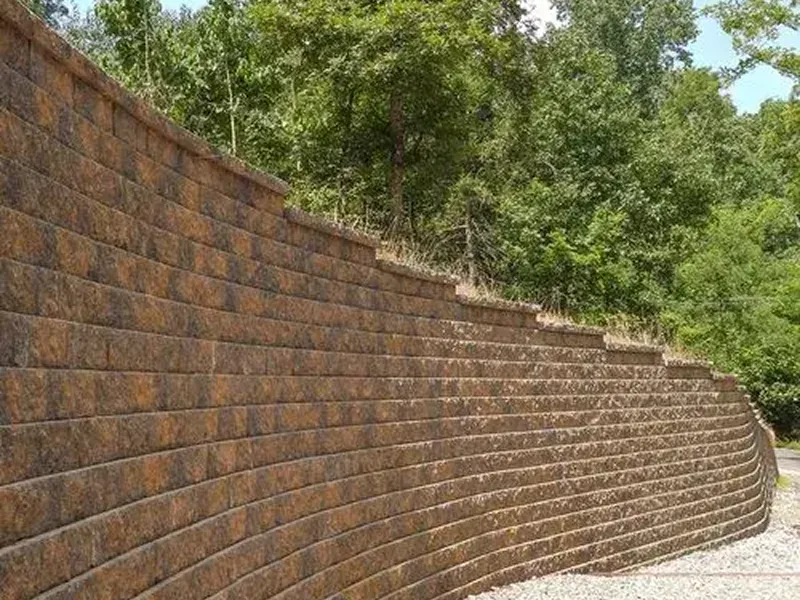 A curved retaining wall made of stacked concrete blocks with grass growing on top, set against a backdrop of green trees.