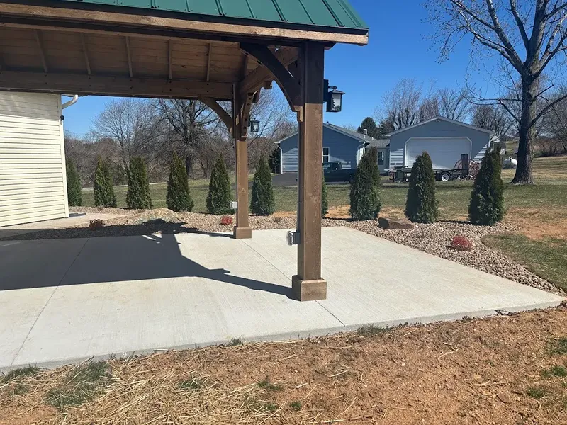A covered patio with wooden posts stands on a concrete slab, bordered by a row of evergreen shrubs and decorative rocks in a suburban backyard.