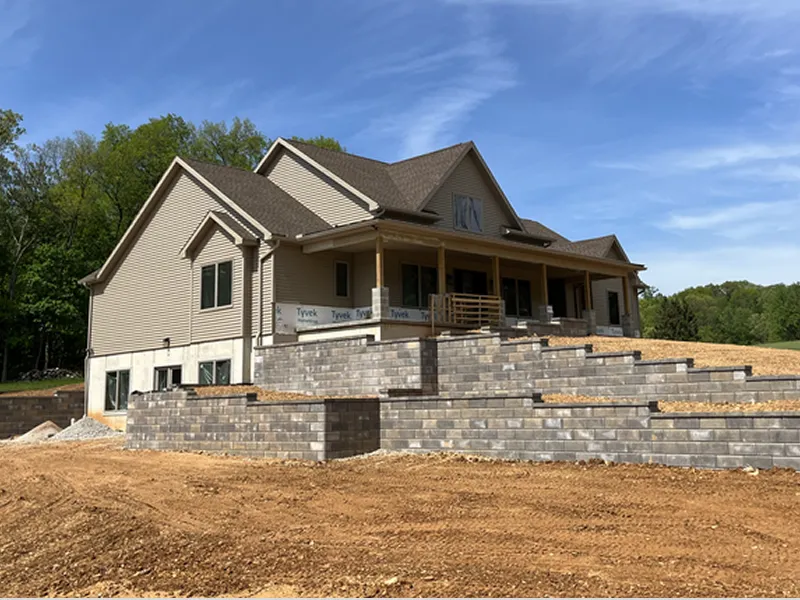 A newly constructed two-story house with a large porch and tiered stone retaining walls, set on a dirt lot with trees in the background under a clear sky.