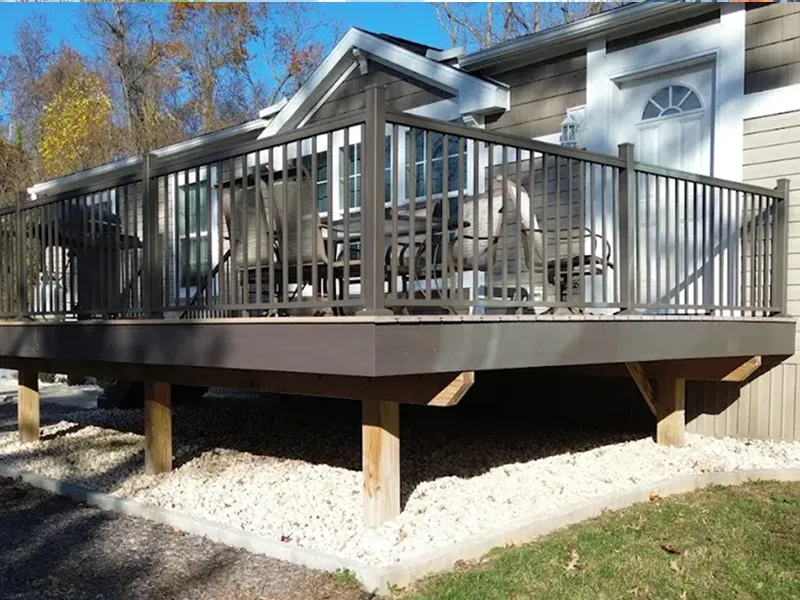 A raised wooden deck with metal railings and outdoor furniture is attached to a house, supported by wooden posts over gravel ground.