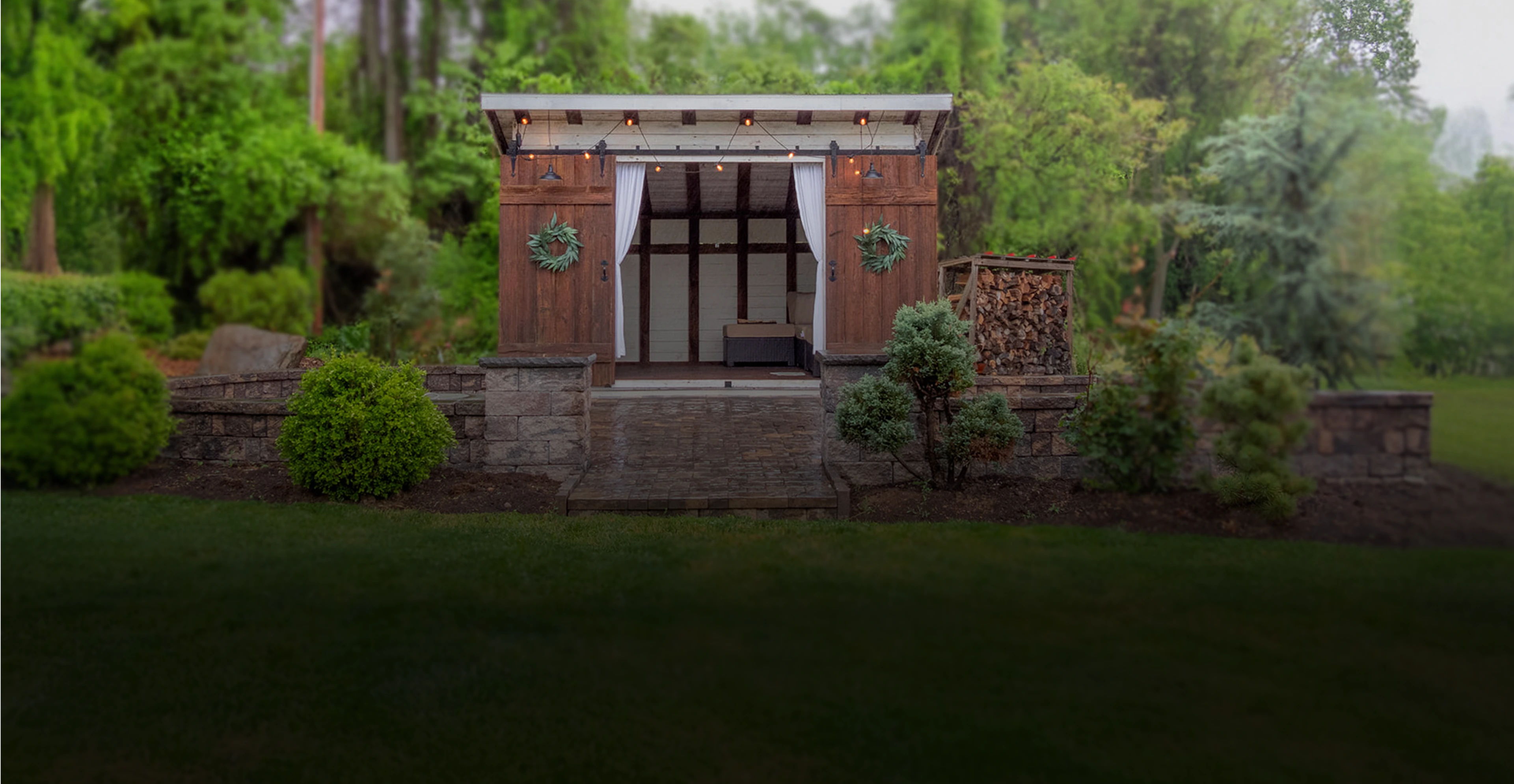 A small wooden garden shed with a tin roof, string lights, and two wreaths, surrounded by greenery and stacked firewood on the right side.