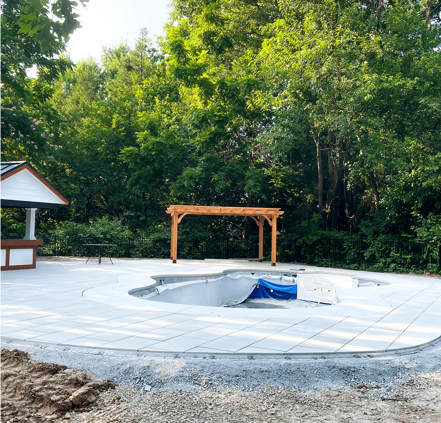 An empty swimming pool under construction, surrounded by newly installed white tiles, with a wooden pergola and dense green trees in the background.