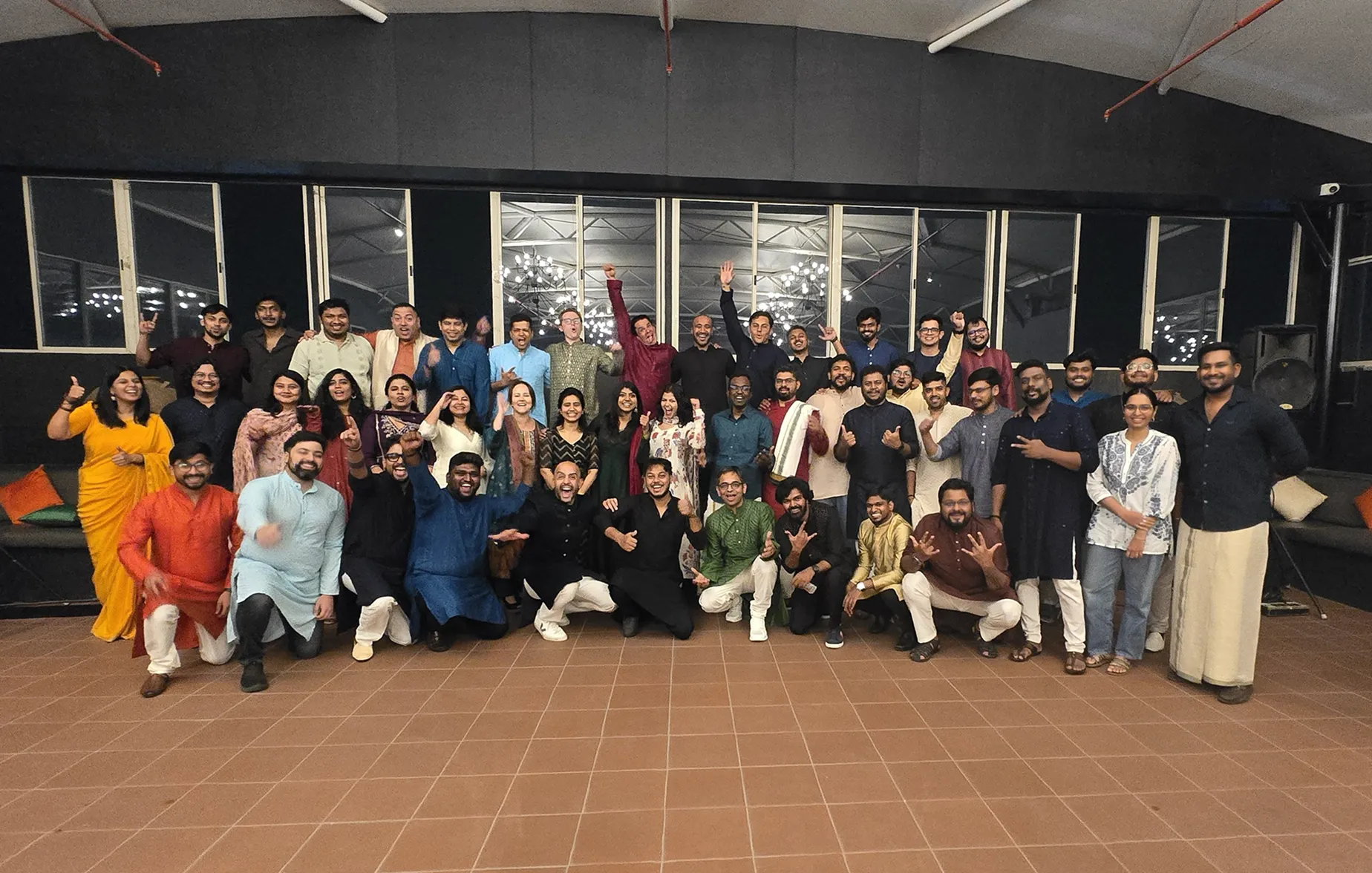 Large group of people posing indoors on tiled floor, many smiling and making celebratory gestures.