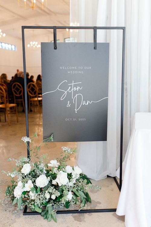 Black 4ft rectangular sign holder for black welcome sign with two leather straps and floral arrangement in bottom left corner