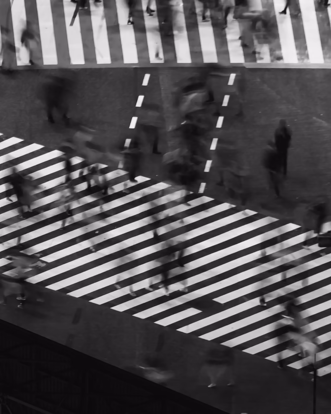 Blurred black and white photo of people crossing a busy street intersection with bold white crosswalk lines.