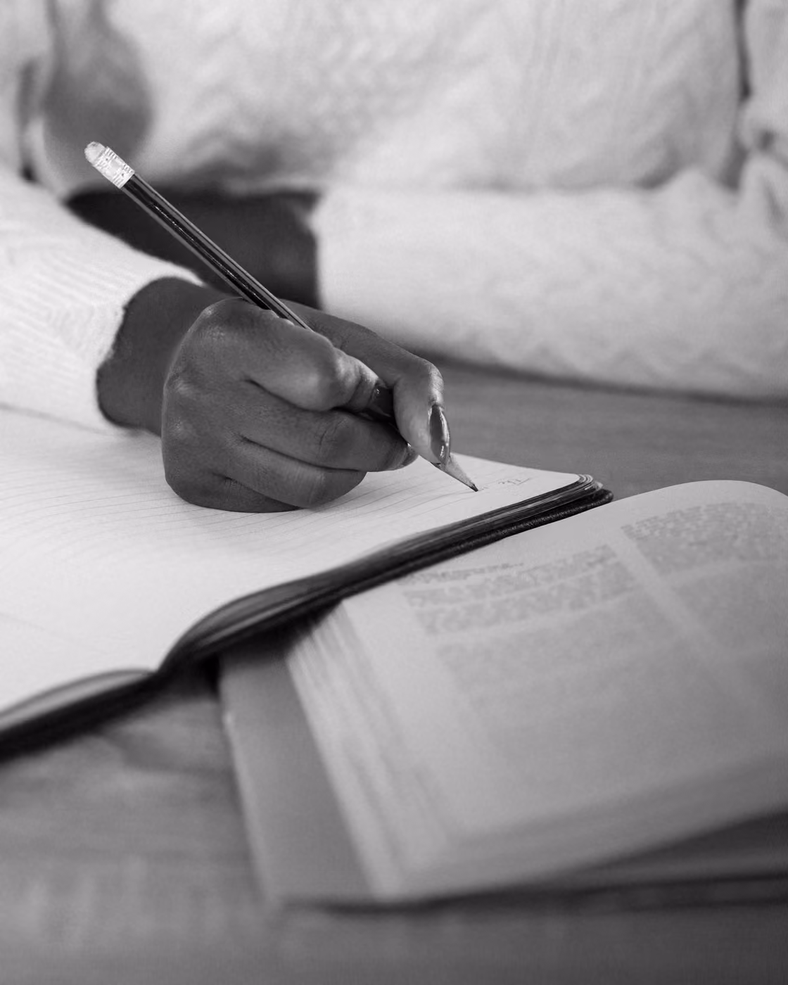 Person writing with a pencil in a notebook, with an open book beside them on a wooden table.