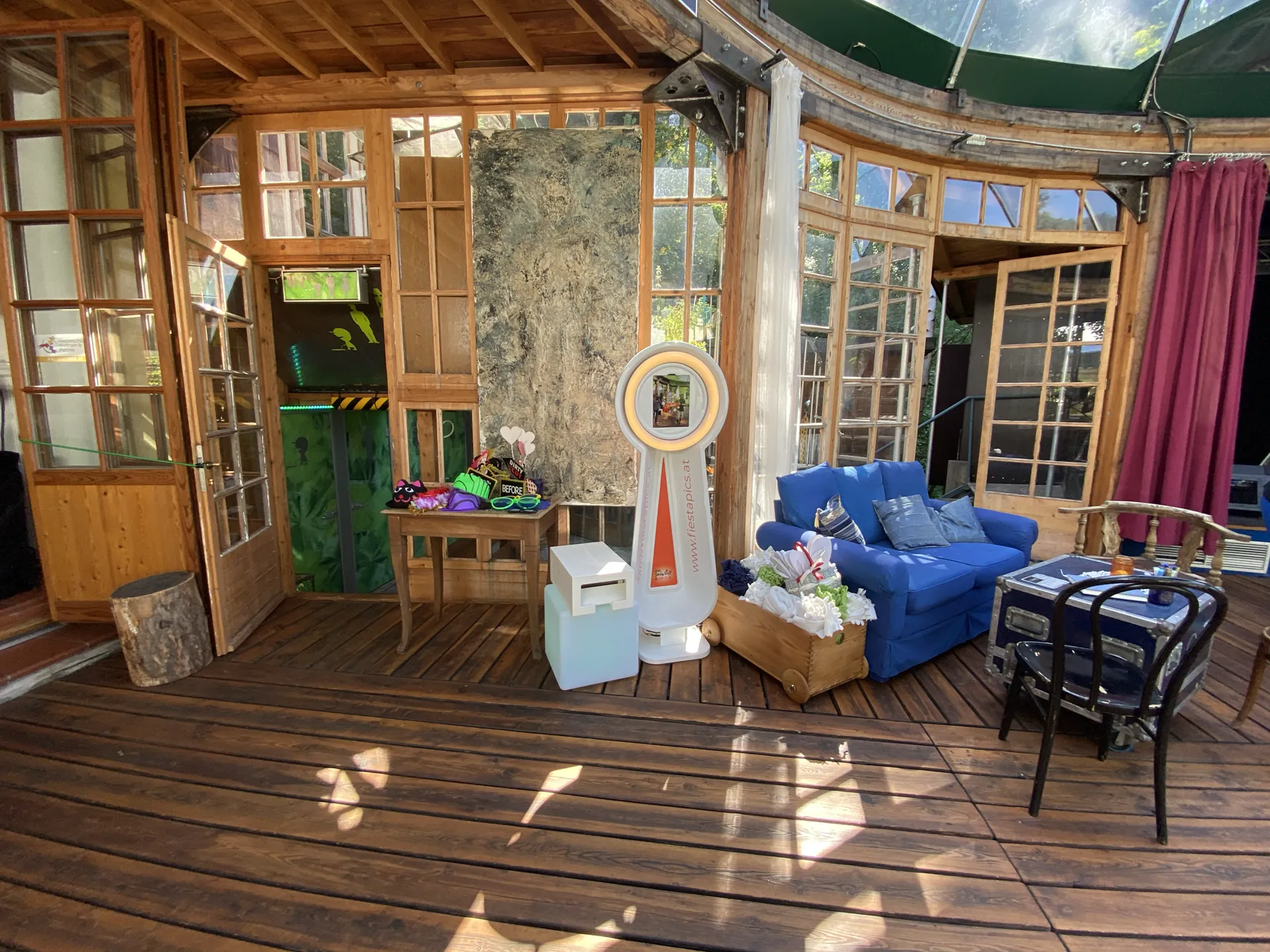 Videobooth in einem Wintergarten mit Holzdecke, daneben ein Tisch mit Verkleidungsutensilien und eine Sitzecke
