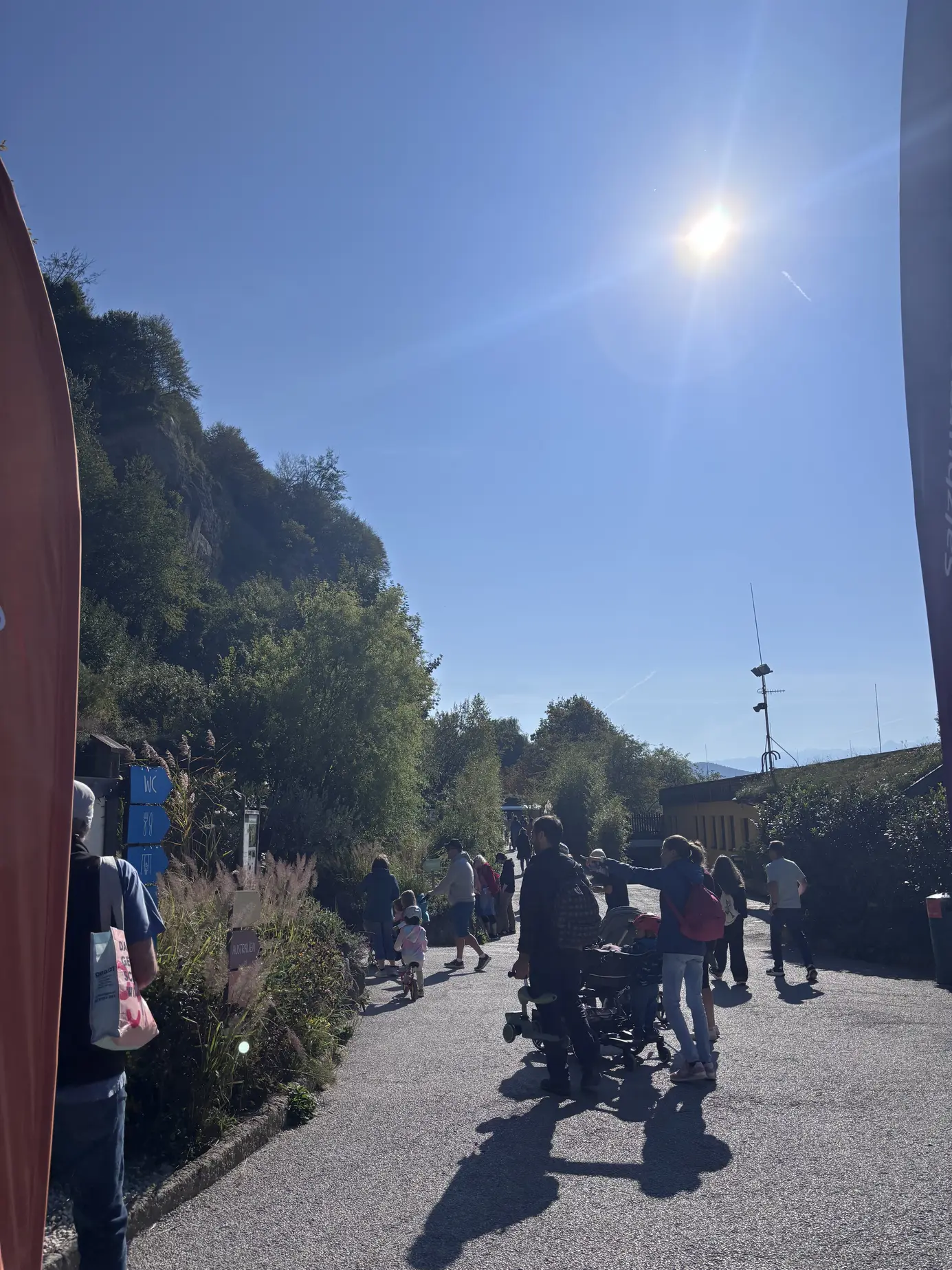 Besucher mit Kinderwagen auf einem Weg im Zoo Salzburg Hellbrunn bei Sonnenschein, links der Hellbrunner Berg