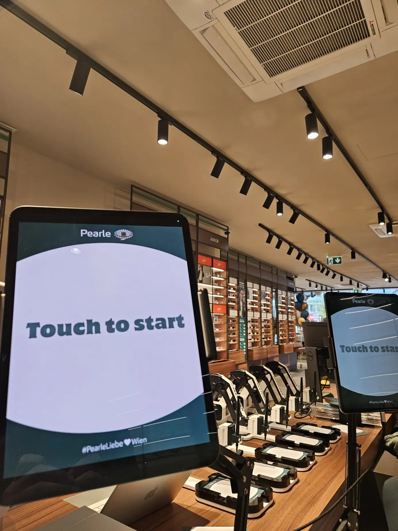 Fotostation des AI Sketchers mit „Touch to start“ im Pearle-Design, dahinter die Roboterarme und die Brillenregale der Filiale