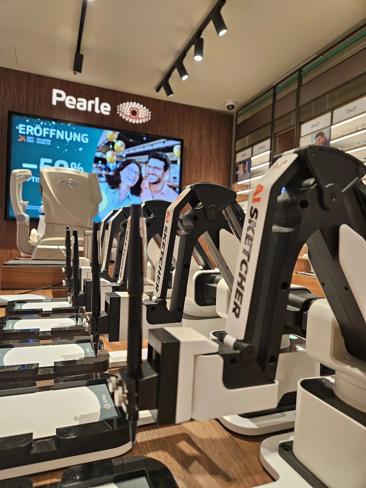 Fünf Roboterarme des AI Sketchers auf einem Holztisch in der Pearle-Filiale, im Hintergrund das Pearle-Logo und der Bildschirm zur Eröffnung