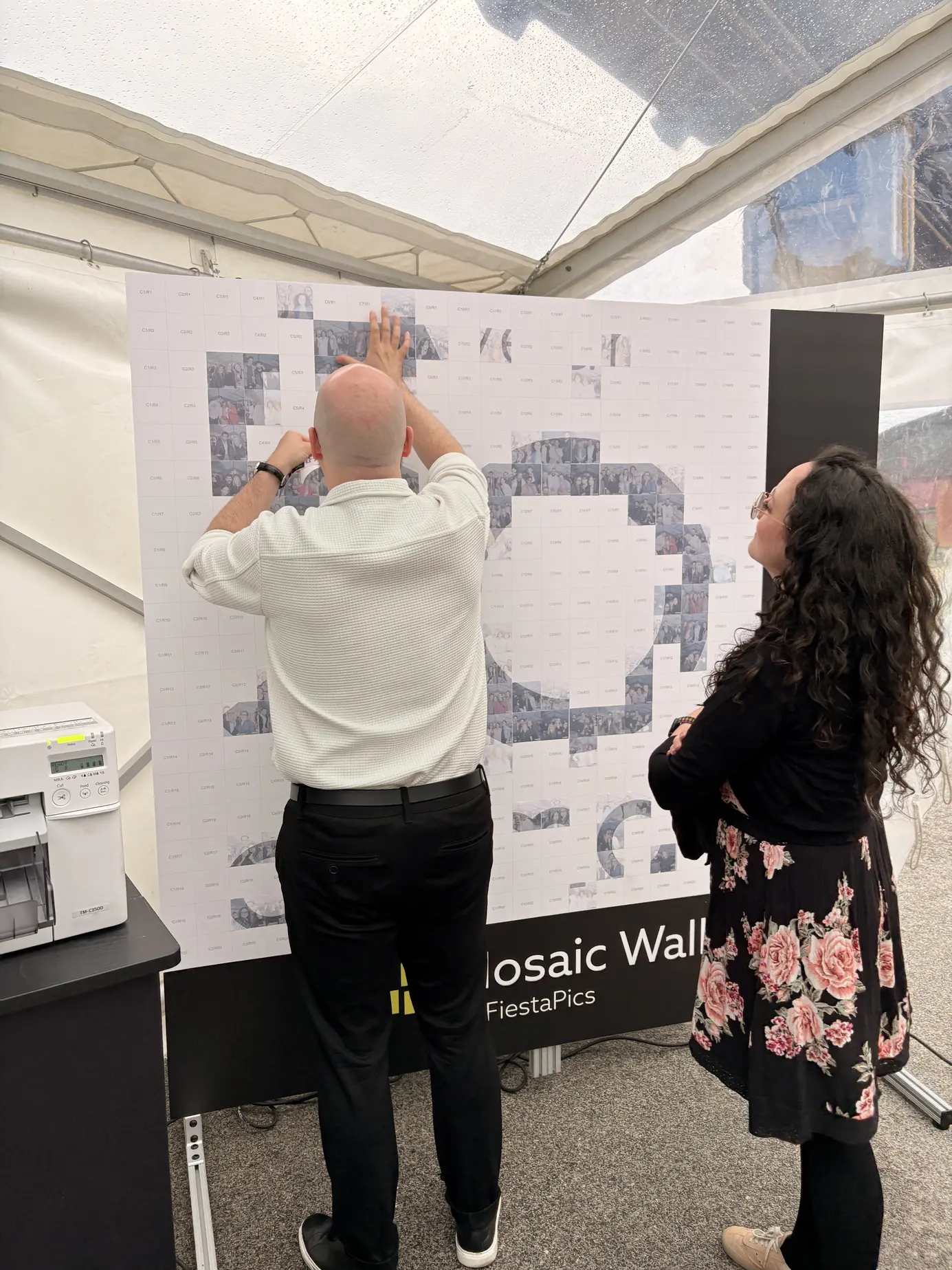 Ein Gast klebt eine Fotokachel auf die Photo Mosaic Wall von FiestaPics im Festzelt, auf der Wand entsteht die Zahl 50, daneben steht der Stickerdrucker