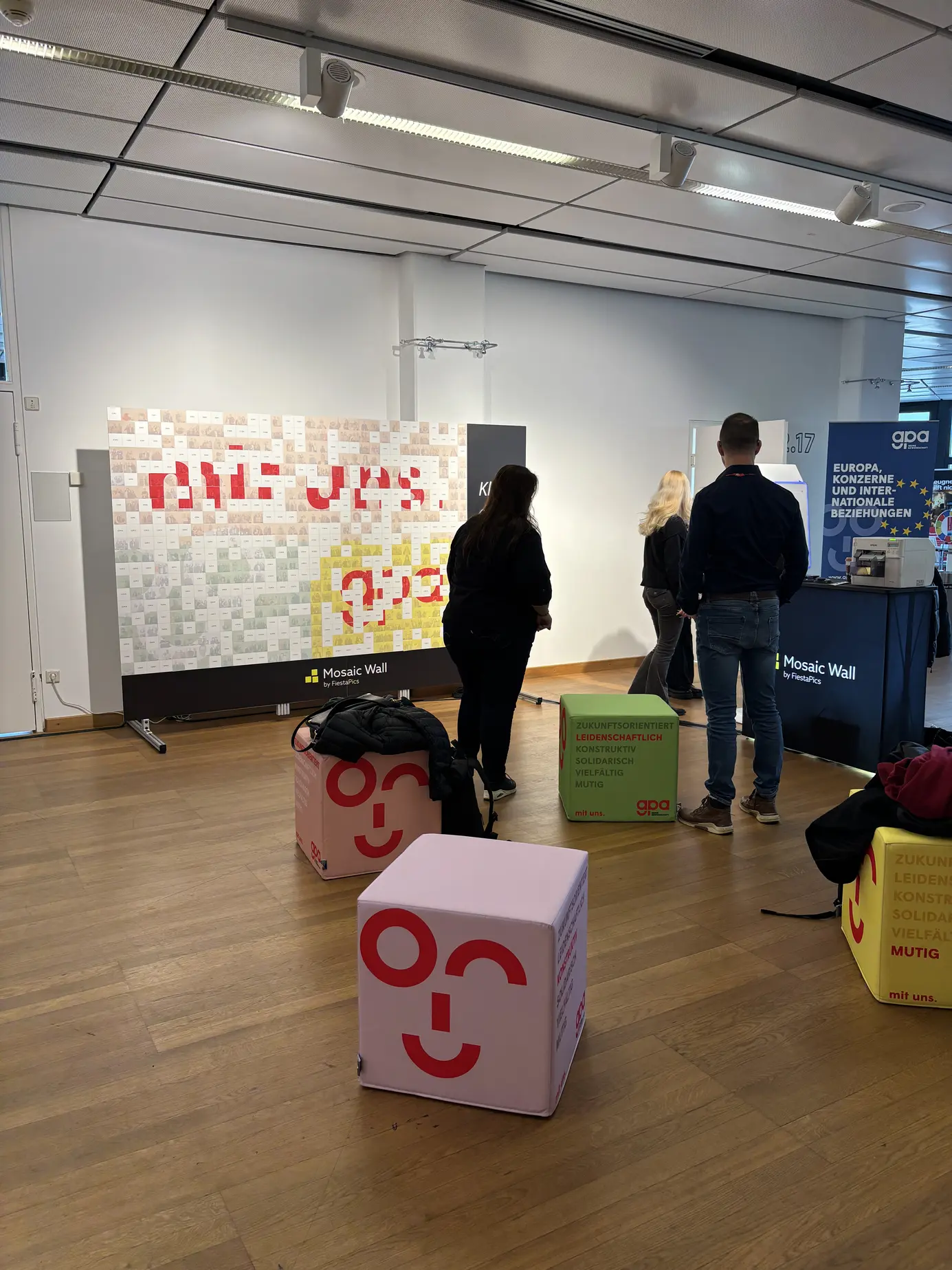 Raum mit der Mosaic Wall im Austria Center Vienna, davor die Sitzwürfel der GPA mit Logo-Gesicht, Besucher an der Wand