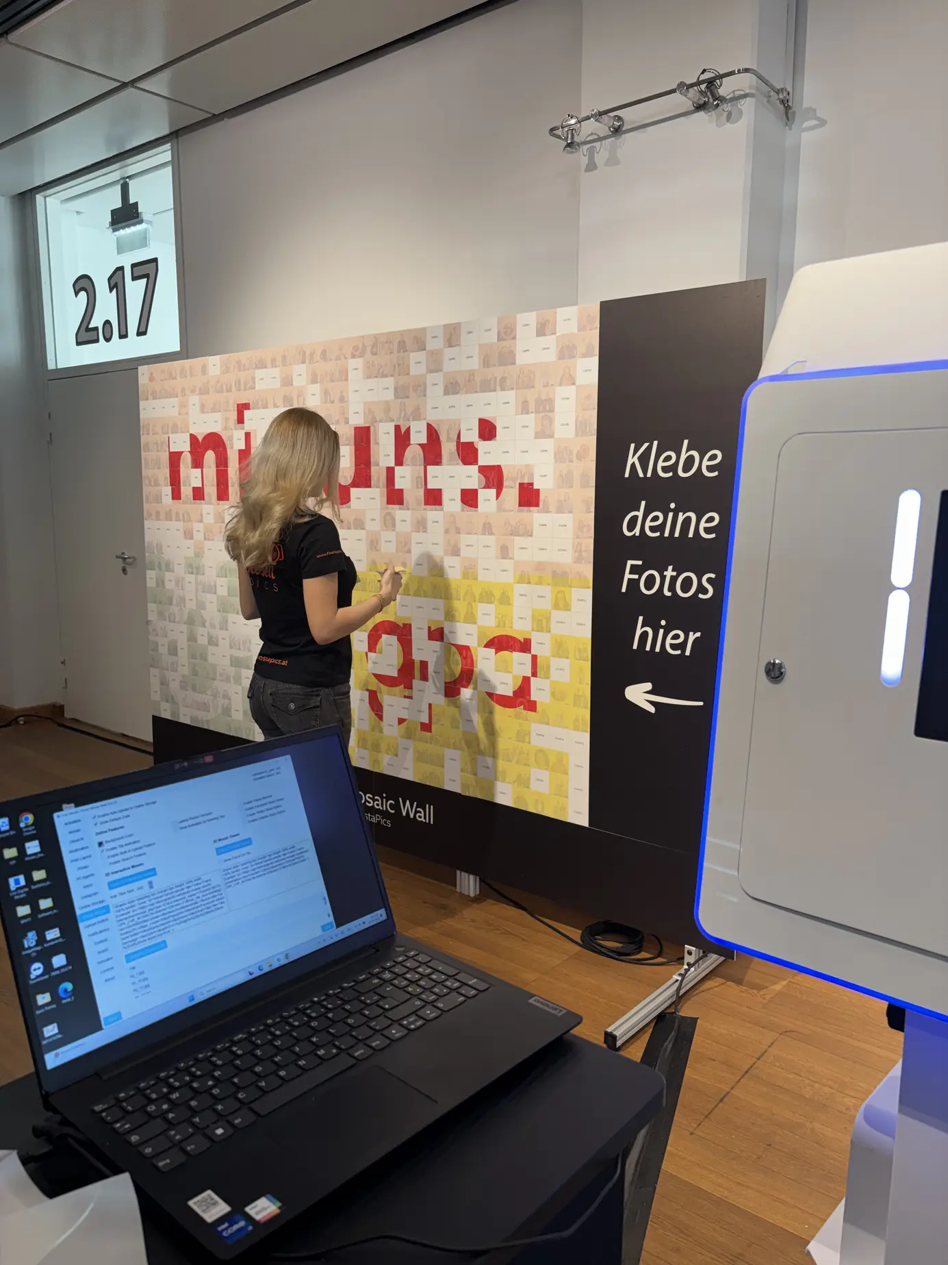 Halb fertige Mosaic Wall mit roten Buchstaben, im Vordergrund Laptop und Fotobox, eine Mitarbeiterin klebt Kacheln