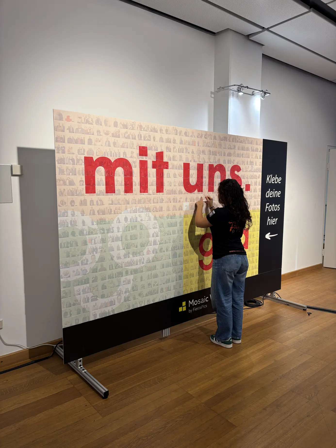 Photo Mosaic Wall von FiestaPics beim Bundesforum der GPA im Austria Center Vienna: Der Schriftzug „mit uns.“ und das GPA-Logo aus hunderten Fotokacheln, eine Mitarbeiterin klebt die letzten Kacheln