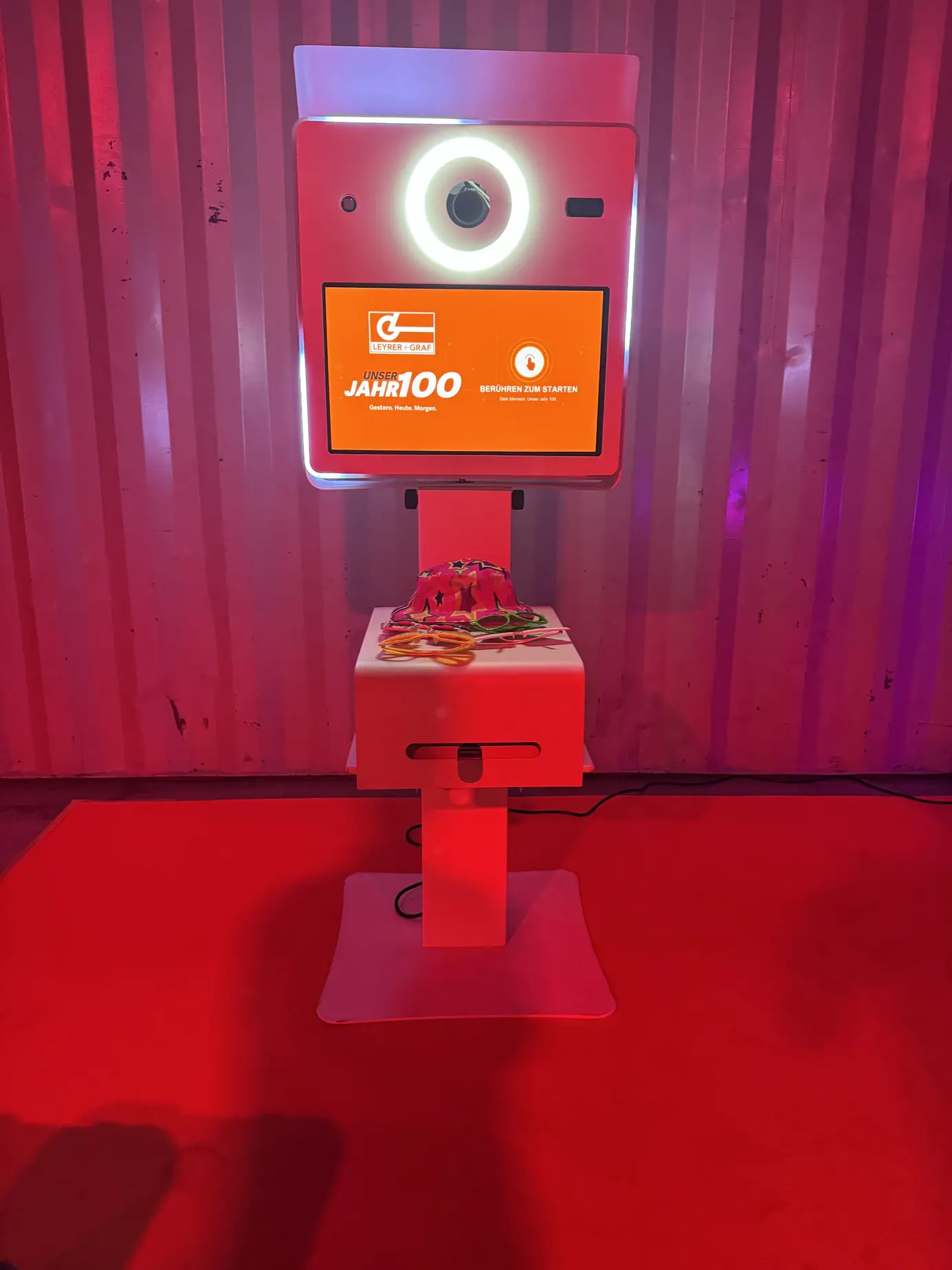 Fotobox Classic mit orangem Jubiläums-Startbildschirm zum 100er von Leyrer + Graf und Fotogadgets auf der Ablage