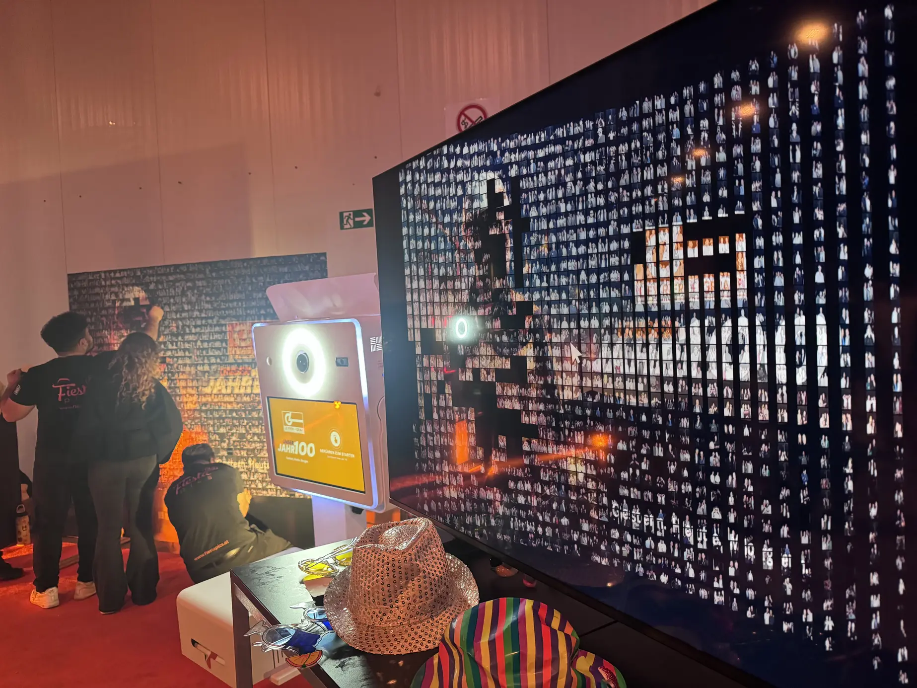 Digitales Live-Mosaik auf großem Bildschirm, dahinter Gäste an der physischen Mosaic Wall und eine Fotobox