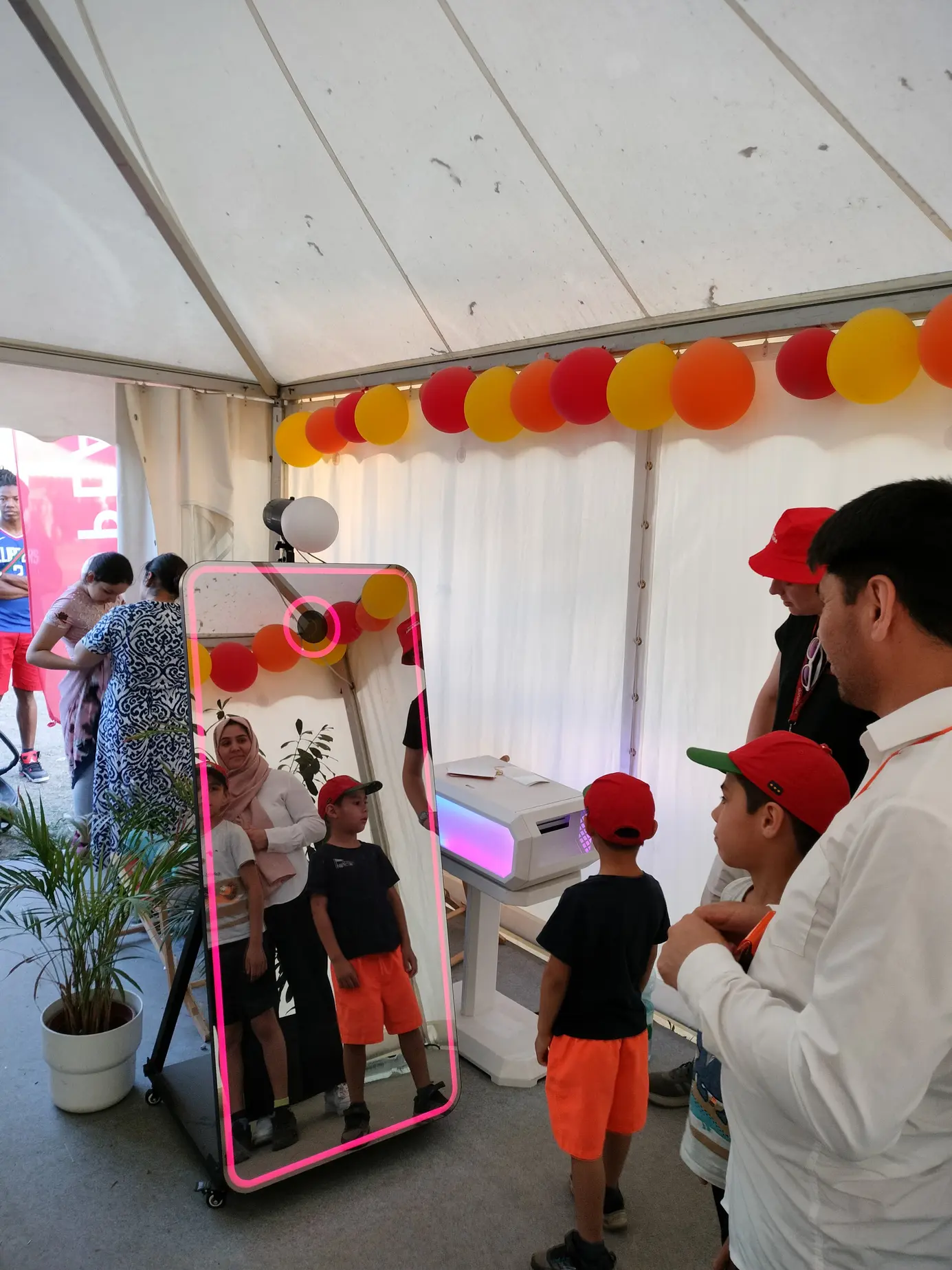 AI Magic Mirror X am Stand von Allwyn im Zelt am Donauinselfest 2025, Kinder mit roten Kappen spiegeln sich im Display, daneben der Fotodrucker