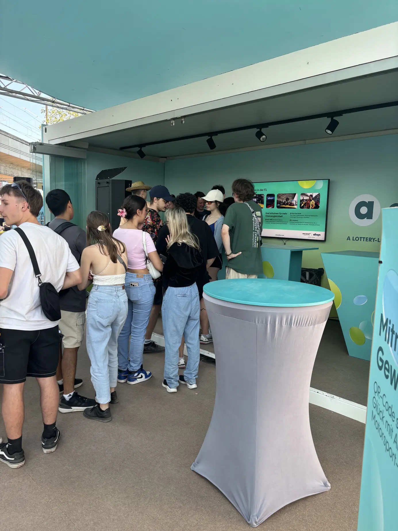Junge Festivalgäste stehen am mintgrünen Stand von Allwyn am Donauinselfest Schlange, am Bildschirm laufen Fotos