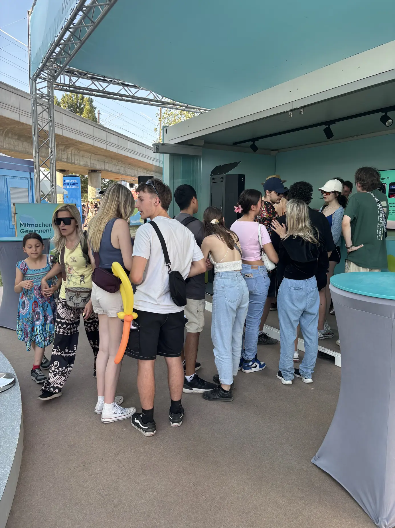 Besucher vor dem Allwyn-Stand am Donauinselfest mit Mitmachen-und-Gewinnen-Aufsteller, im Hintergrund die Brücke über die Donauinsel