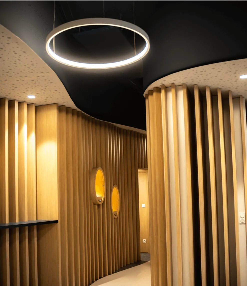 Agencement intérieur bois sur mesure : espace feutré aux cloisons courbes en tasseaux de bois, grande suspension circulaire et niches ovales éclairées chaudement.