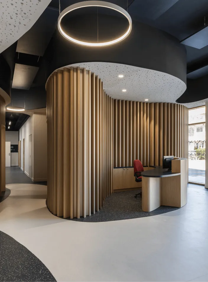 Agencement intérieur bois sur mesure : espace d'accueil aux cloisons courbes en tasseaux de bois clair, suspension lumineuse circulaire et banque arrondie.