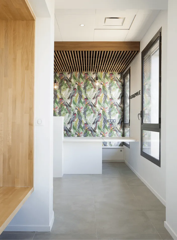 Aménagement en menuiserie sur mesure : coin bureau au plafond en tasseaux de bois, papier peint tropical à perroquets et fenêtre grise.