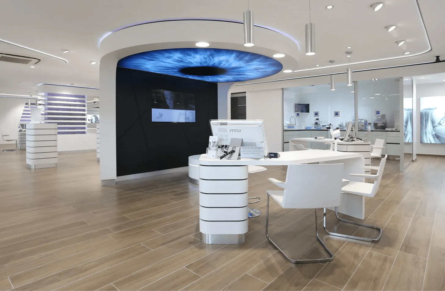 Mobilier et agencement bois sur mesure : showroom d'optique moderne avec comptoirs blancs, écran circulaire lumineux figurant un œil, sol en parquet et chaises design.