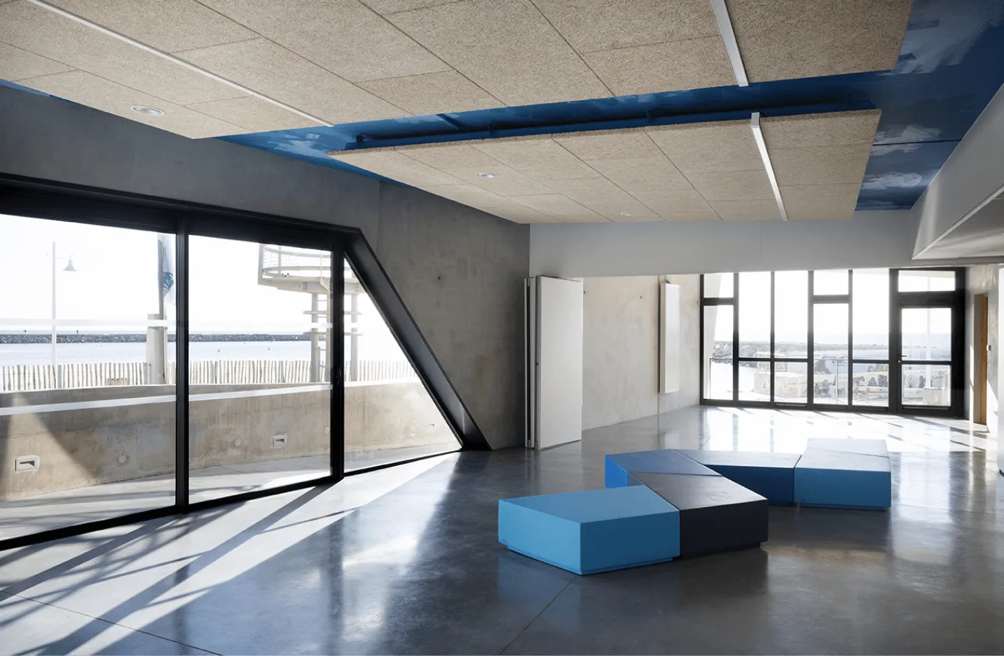 Agencement sur mesure en menuiserie bois : vaste salle béton aux larges baies vitrées, plafond à poutre bleue et assises modulaires.