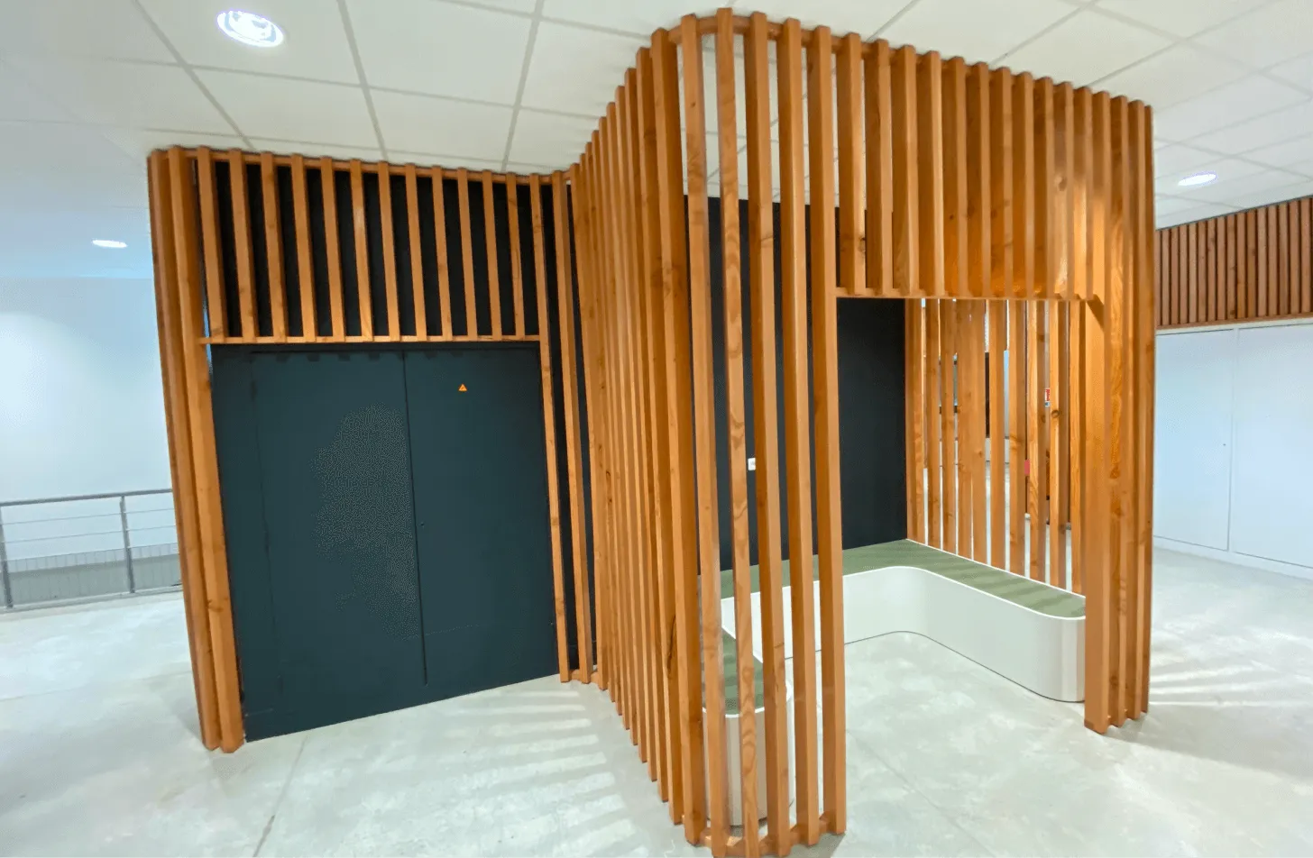 Agencement intérieur bois sur mesure : hall avec habillage sculptural en tasseaux de bois verticaux entourant des portes d'ascenseur sombres, sol clair et plafond à dalles.