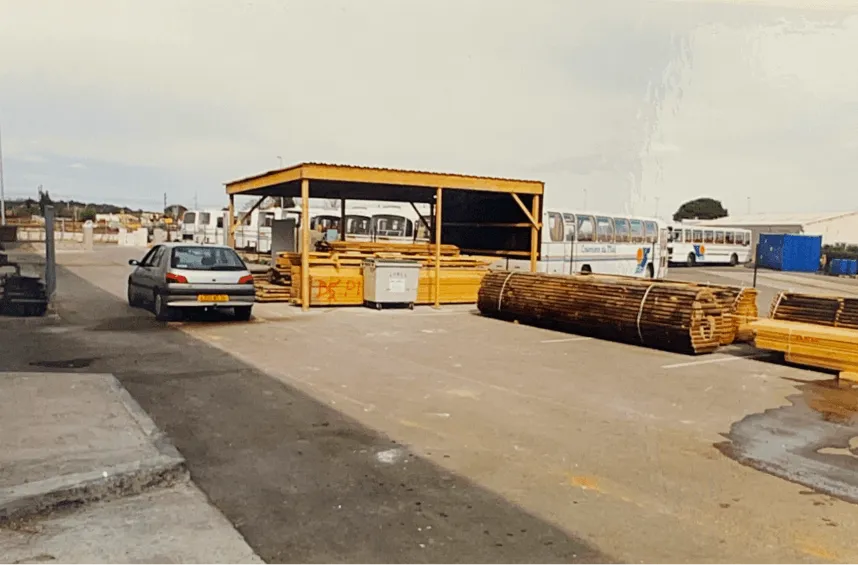 Atelier et parc à bois d'une menuiserie : cour de stockage avec auvent abritant planches et bottes de bois, atelier de menuiserie à Montpellier, véhicules stationnés.