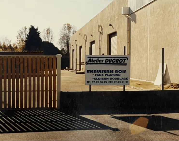 Menuiserie extérieure et bardage bois sur mesure : façade des Ateliers Ducrot, menuiserie bois sur mesure à Baillargues, panneau enseigne, portail en bois et ciel bleu ensoleillé.