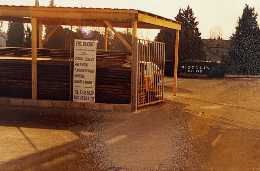 Atelier et parc à bois d'une menuiserie : auvent abritant les stocks de bois de l'atelier de menuiserie de Saint-Clément-de-Rivière, portail métallique et enseigne Ducrot.