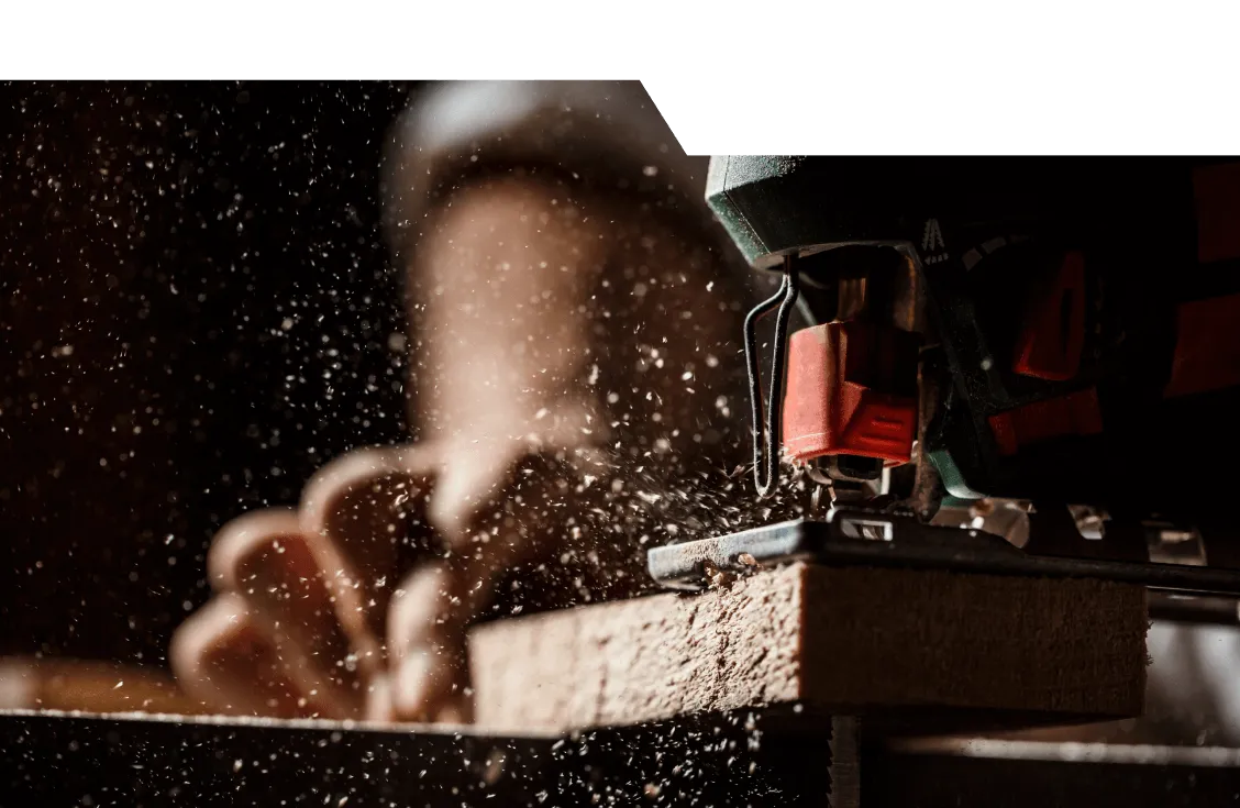 Atelier de menuiserie : gros plan d'une scie sauteuse découpant une planche de bois, avec projection de sciure dans l'atelier sombre.